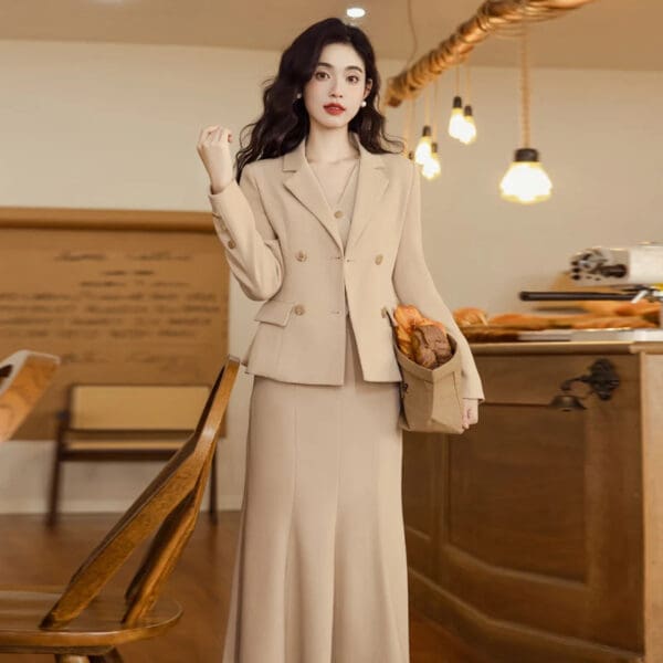 A woman in a beige suit standing next to a wooden table.